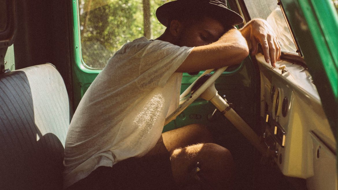 Rapaz de chapeu, t-shirt e calções numa camioneta antiga, com sono e com a cabeça no volante